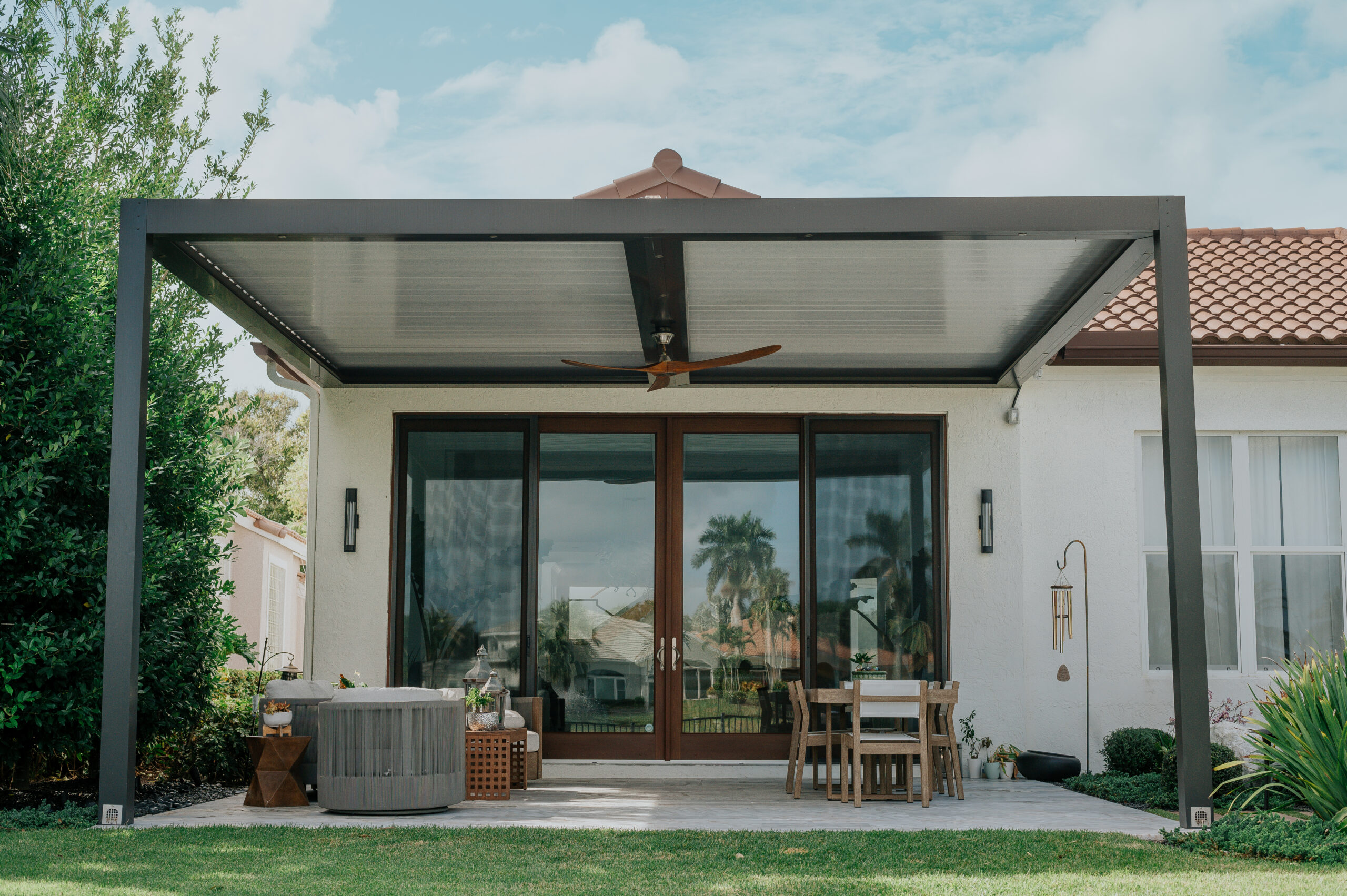 A dark grey attached motorized louvered pergola over a paved patio with a teak dining set and a hot tub, against a white house with a tile roof.