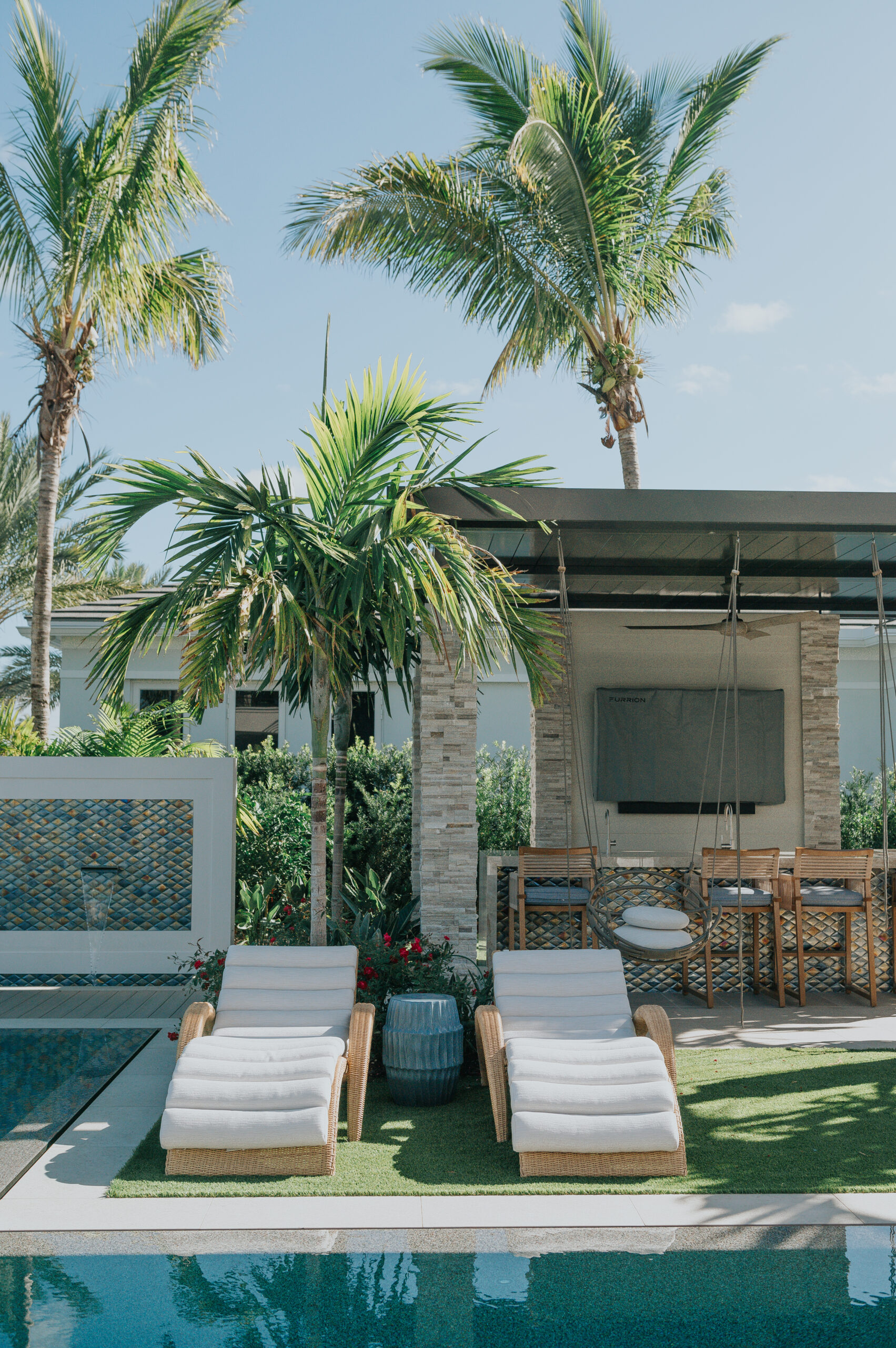 A poolside cabana bar under a dark pergola with an outdoor television, teak bar stools and sun loungers, beside a tiled water feature and palms.