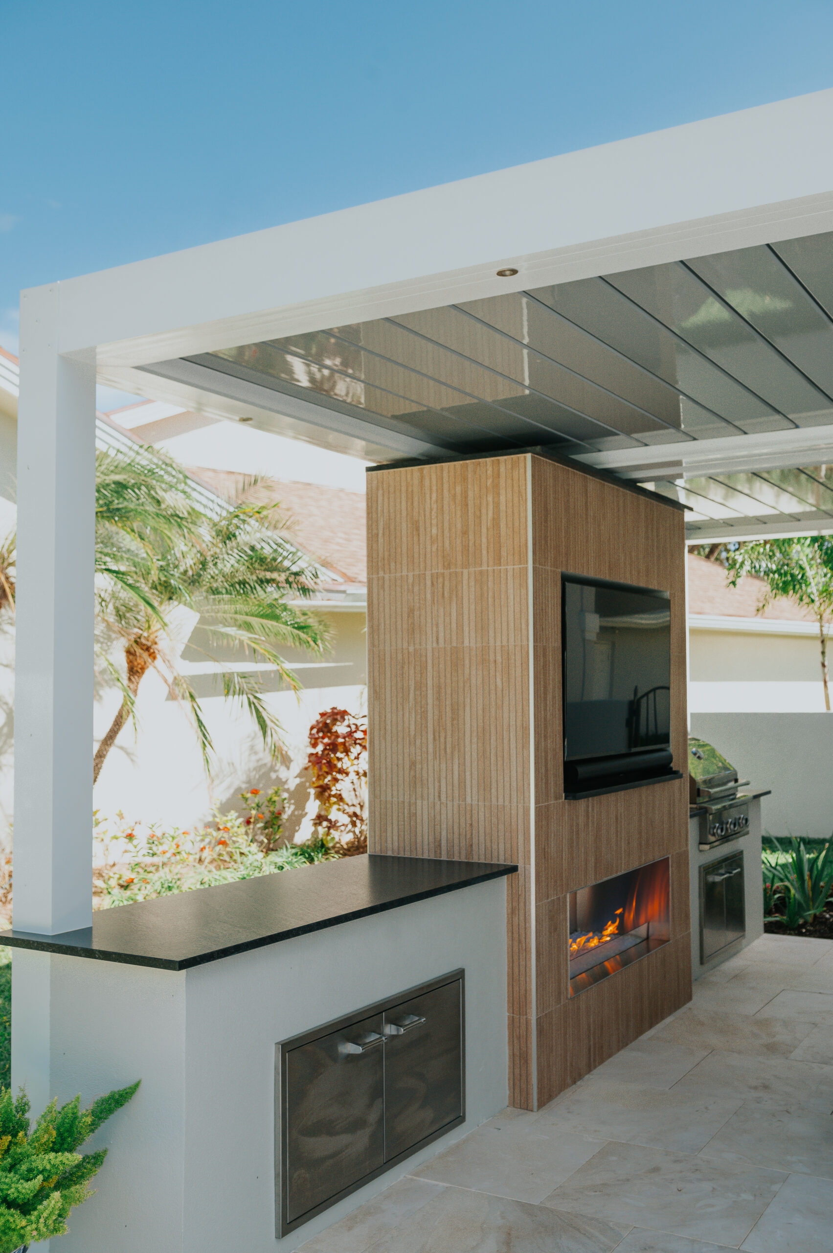 An outdoor kitchen beneath a white motorized louvered roof, with a tiled feature wall carrying a television above a linear gas fireplace, a granite counter, built-in grill and stainless cabinet doors.