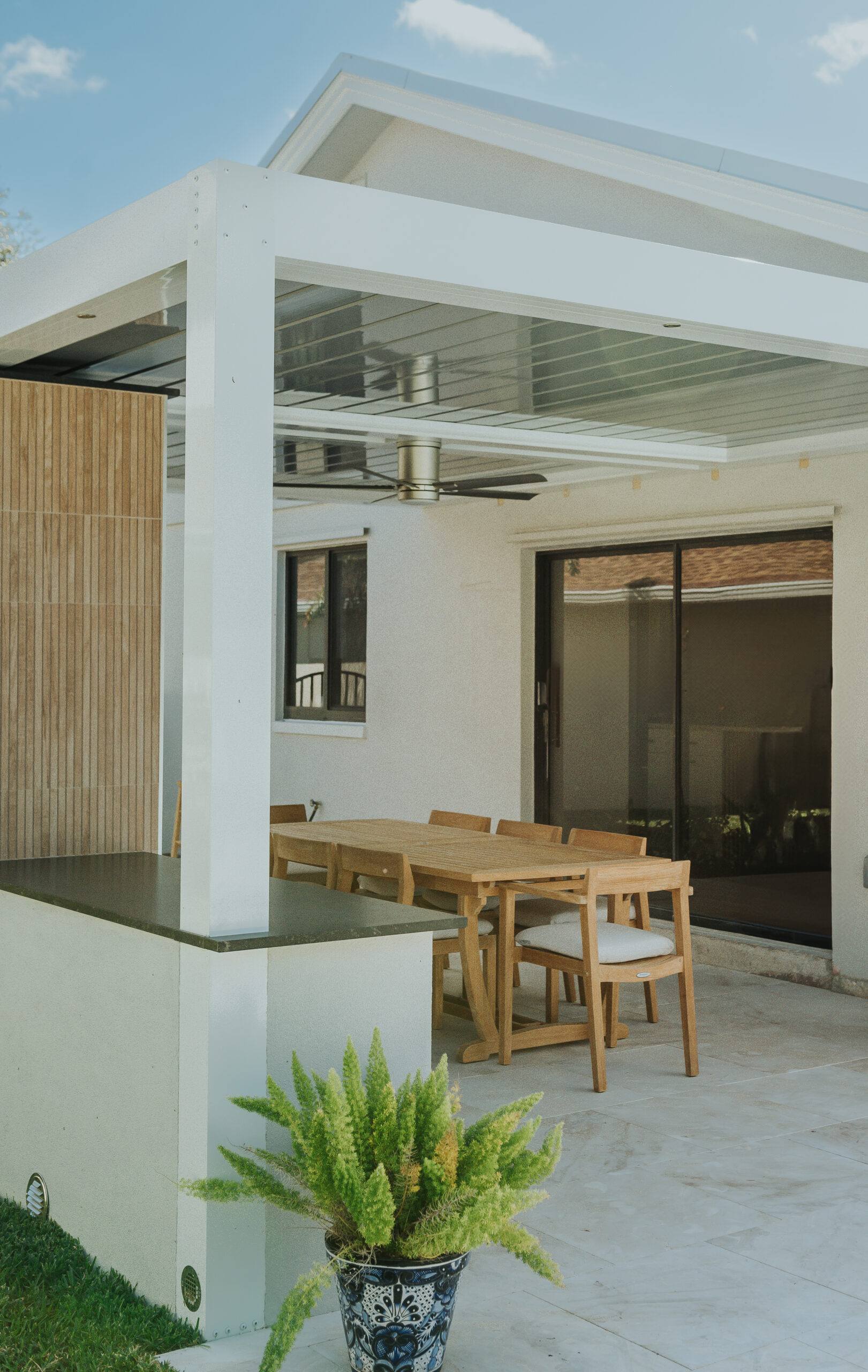 A white attached motorized louvered pergola over a teak dining table and chairs beside an outdoor counter, against a stucco house with sliding glass doors.
