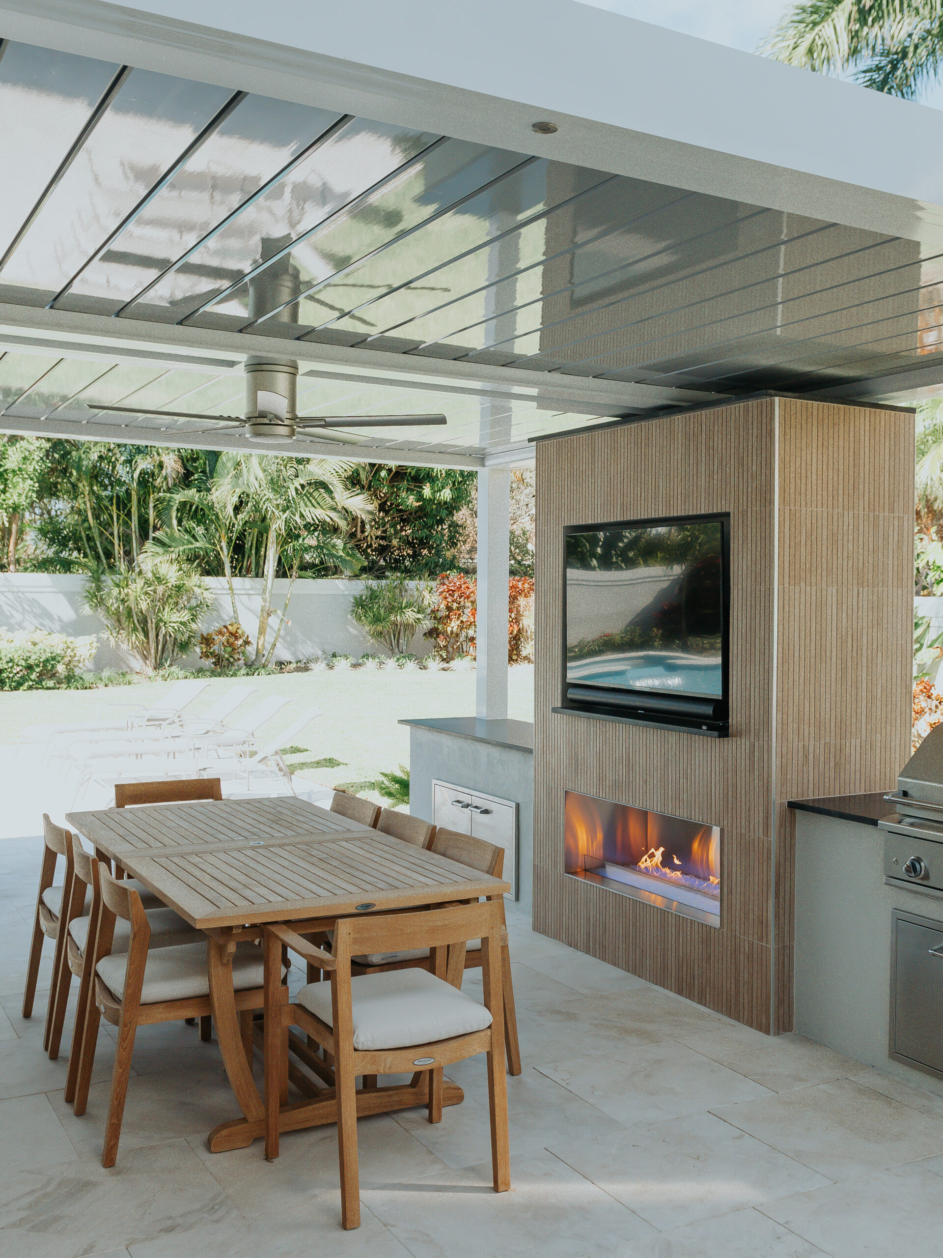 A white motorized louvered roof over an outdoor dining table, with a tiled wall carrying a television above a linear fireplace, a built-in grill alongside, and a pool beyond.