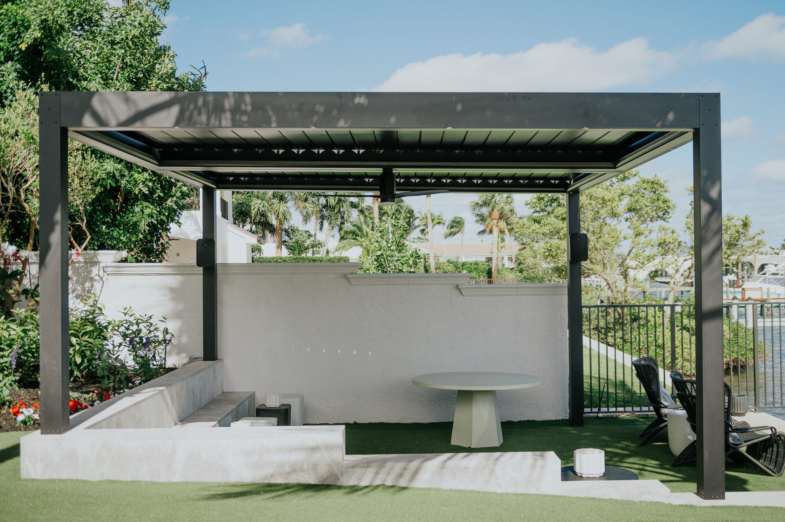 A dark freestanding motorized louvered pergola on artificial turf beside a waterway, with the louvers open to the sky, a round table and lounge chairs.