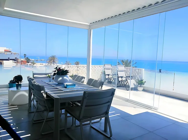 A frameless sliding glass enclosure around a terrace with a dining table, opening onto a sea view.